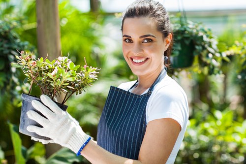 A smiling woman in a gardening apron and white gloves is holding a potted plant with green and purple foliage, standing in a lush garden with a variety of shrubs, flowering plants, and a mature tree in the background. The garden features well-maintained grass, gravel pathways, and wooden garden structures, suggesting a thoughtfully landscaped outdoor space typical of residential gardens in Blackheath. Bright natural light indicates a clear day, highlighting the vibrant green tones and natural textures of the plants and garden surfaces. This scene reflects professional outdoor maintenance and gardening services, focusing on plant care, garden design, and overall landscape enhancement relevant to local outdoor spaces in the Blackheath area.