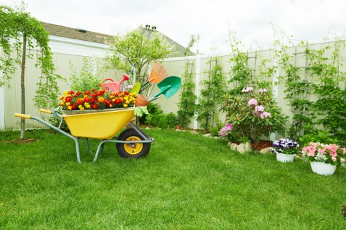 A neatly maintained suburban backyard garden featuring a well-kept green lawn with lush grass. In the center, a bright yellow garden wheelbarrow is filled with colourful gardening tools and potted flowers, indicating active planting or garden maintenance. To the right, a flower bed edged with red bricks displays a variety of blooming pink and purple flowering plants, possibly including rhododendrons or azaleas. The garden is enclosed by a white wooden picket fence, with several climbing plants and vines growing along its surface, adding greenery and privacy. In the background, a small tree with fresh green leaves provides some shade, and the overall scene appears bright and sunny, suggesting fair weather. The layout is simple yet functional, suitable for outdoor gardening activities typical of a Blackheath or South East London garden, with visible elements that highlight lawn care and flower planting services offered by landscapers in the area.