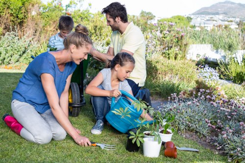 A family garden scene showing a lush, well-maintained lawn with neatly edged flower beds filled with a variety of flowering plants and shrubs. The foreground features a young girl digging in the soil using a trowel, accompanied by an adult woman who is kneeling on the grass, assisting her. Nearby, gardening tools, a watering can, and plant pots are arranged on the lawn, indicating ongoing gardening activities. In the background, a man and another child are engaged in planting or tending to the garden, with dense greenery, small bushes, and blooming flowers creating a vibrant, colorful outdoor environment. The garden is part of an outdoor space with natural lighting under a clear sky, suggesting a bright, pleasant day. This scene highlights typical garden maintenance and planting activities that home gardening services like those from Landscapers Blackheath might support, with a focus on well-kept lawns, flower beds, and planting areas in a suburban setting close to Blackheath, London, with the backdrop hinting at scenic views and a peaceful residential backdrop.