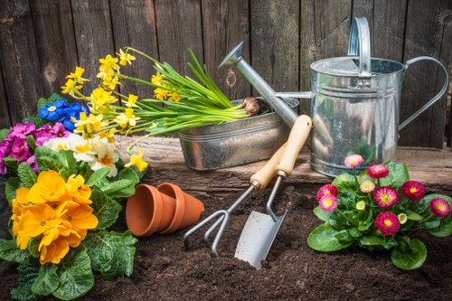 A garden scene featuring a freshly prepared flower bed with rich, dark soil, surrounded by colourful flowering plants including yellow daffodils, pink primroses, and purple pansies. In the background, a weathered wooden fence encloses the outdoor space, which appears to be part of a landscaped backyard in Blackheath, UK. Gardening tools such as a watering can, a small hand rake, and a trowel rest on the soil, alongside a pair of empty terracotta pots and a metal container filled with vibrant spring flowers, ready for planting or tending. The scene is illuminated by natural daylight, suggesting a mild, bright day suitable for outdoor gardening activities, aligning with professional gardening and landscaping practices typical in the local area.