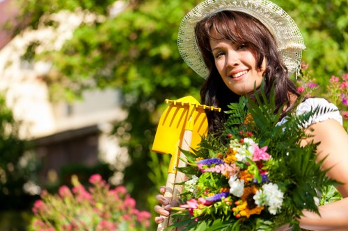 A smiling woman in a white summer dress and wide-brimmed straw hat is standing in a well-maintained garden, holding a colorful bouquet of flowers and a yellow garden rake. The garden features a lush, green lawn with a dense hedge in the background, alongside a variety of flowering plants and shrubs that add vibrant pink, purple, and white hues. Sunlight filters through the trees, illuminating the scene with natural light, creating a bright and inviting outdoor environment. The garden area appears neatly bordered, with natural textures of grass, foliage, and flowering plants clearly visible, indicating ongoing gardening or landscaping care by Landscapers Blackheath. The setting reflects a typical front or back garden space in Blackheath, showcasing a combination of cultivated planting and natural outdoor elements suitable for professional gardening and landscaping services.