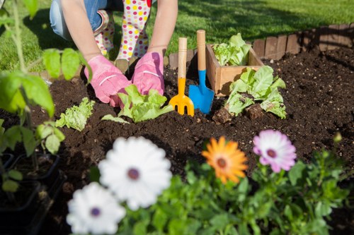 The image shows a garden bed in a landscaped outdoor area, likely within a residential garden in Blackheath, with rich, dark soil prepared for planting. In the foreground, there are flowering plants with white, orange, and purple blooms, providing vibrant colour contrast against the dark soil. Behind these flowers, a person wearing pink gardening gloves and colorful clothing is actively planting or tending to young lettuce or leafy greens, which are arranged in neat rows within the garden bed. The garden includes small wooden plant supports or markers, along with gardening tools such as a small trowel and rake, positioned in the soil beside the planting area. The backdrop features a well-maintained lawn with lush green grass, and a border of a brick or stone edging is visible along the garden bed, contributing to a neat and structured outdoor space. The environment appears sunny and bright, indicating good weather, suitable for gardening activities, and reflecting the professional outdoor landscaping or gardening services provided by Landscapers Blackheath in the local area.