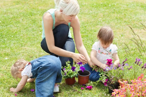 A woman with short blonde hair, wearing a light blue sleeveless top and dark jeans, is kneeling on a well-maintained lawn in a garden in Blackheath. She is planting or tending to potted plants, including purple and pink flowering plants, which are placed on the grass. Two young children, a girl with light brown hair dressed in a white top with floral patterns and a boy in a blue outfit, are engaged in similar gardening activities nearby, with the girl holding a small gardening tool and the boy examining the ground. The garden features a lush, green grassy area with a variety of flowering plants and potted plants arranged around the women and children. The setting appears to be on a clear, sunny day, with natural light illuminating the scene, highlighting the vibrant colors of the flowers and the healthy, dense lawn. This outdoor environment exemplifies typical features of a landscaped backyard garden in Blackheath, suitable for family gardening projects, tended to by expert landscapers like Landscapers Blackheath, who provide professional gardening and outdoor maintenance services in the local area.