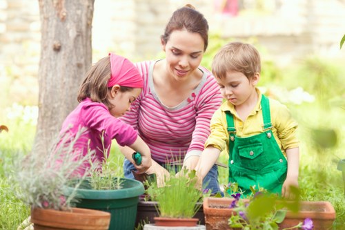 A woman with dark hair tied back and wearing a pink and white striped top is engaged in gardening in an outdoor garden space, accompanied by two young children. One child, wearing a pink headband and pink jacket, is planting or tending to a potted plant, while the other child, dressed in a yellow shirt with green overalls, observes close by. The garden features a variety of potted plants and flowers, with some pots made of terracotta and others plastic, arranged on a wooden surface or garden bed. The background includes a mature tree with textured bark and a blurred stone or brick wall, indicating a landscaped backyard environment. Natural sunlight illuminates the scene, highlighting the lush greenery, vibrant flower colors, and the active, engaged atmosphere of outdoor gardening, reflecting typical garden maintenance and planting activities as offered by local Blackheath landscape gardeners.