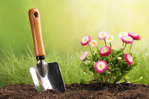 A close-up view of a garden bed featuring rich, dark soil with a small hand trowel partially inserted into the ground, its wooden handle clearly visible. To the right, there is a vibrant cluster of pink and white flowers, possibly primroses or similar, with lush green leaves surrounding them. The background is softly blurred, showing a well-maintained grassy lawn and hints of natural sunlight, indicating a bright, clear day. This outdoor scene exemplifies typical landscaping elements found in a UK garden, with neatly cultivated flower beds and a clean, tidy lawn area, suggesting attention from professional gardening services such as those by Landscapers Blackheath. The setting appears to be part of a landscaped front garden or backyard, with natural tones of soil, flowers, and grass blending seamlessly to create a welcoming outdoor space suitable for ongoing gardening and lawn care activities.